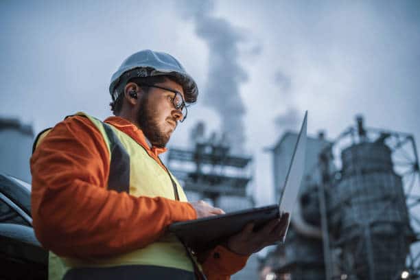 A shot of an young engineer wearing a helmet and using a laptop and hands free device during his night shirt in the oil rafinery. Engineering concept.